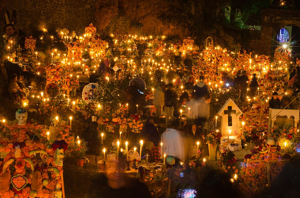 Scène de cimetière éclairée à la bougie, avec des fleurs colorées et des gens rassemblés, créant une atmosphère chaleureuse et sereine sous un ciel sombre.