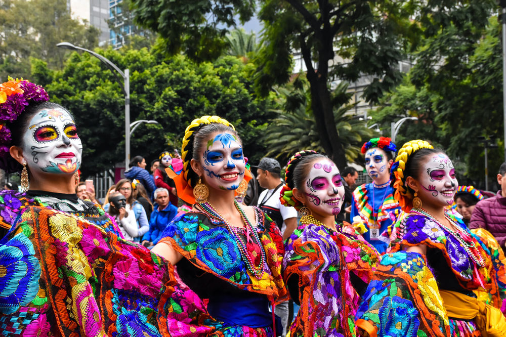 Des personnes en costumes colorés du Jour des morts, le visage peint, défilent joyeusement dans un parc aux arbres verdoyants, devant une foule rassemblée.
