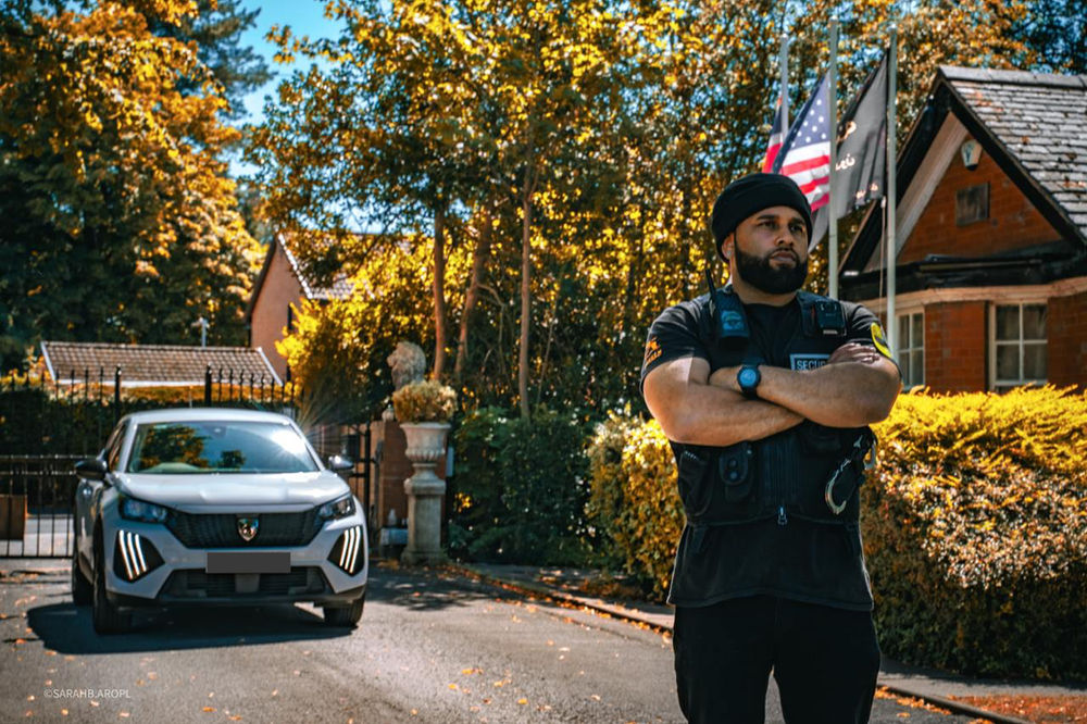 Un agent de sécurité se tient bras croisés près d’un SUV, devant une maison close de grilles, avec des arbres d’automne et des drapeaux. Un soleil vif projette des ombres.