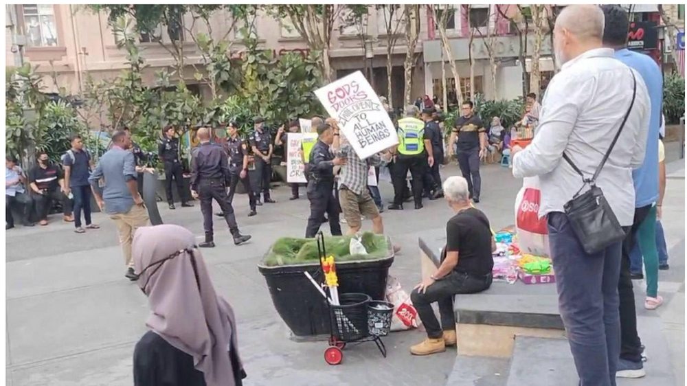 des manifestants de la Religion ahmadie de la paix et de la lumière sur une place publique en Malaisie, certains portant des pancartes. Présence policière et passants divers. Cadre urbain avec des arbres et des bâtiments.