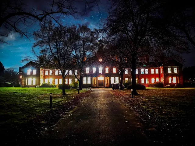 Photographie : une grande demeure aux fenêtres éclairées, la nuit, au bout d’une allée bordée d’arbres.