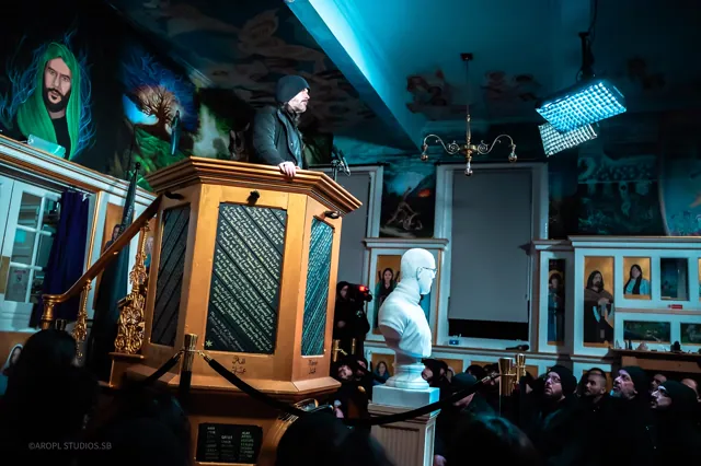 Photographie : un homme s’adresse à une assemblée depuis une chaire de bois sculpté, dans une salle aux murs peints.