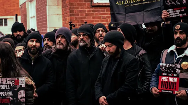Photographie : des manifestants en bonnets noirs, devant un bâtiment de briques, tiennent des pancartes dont l’une porte « Free them now! ».