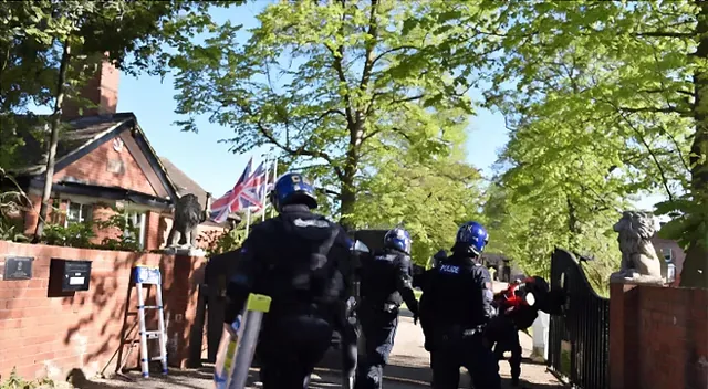 Capture d’écran : des policiers casqués franchissent le portail d’une propriété en briques, bordée d’arbres.